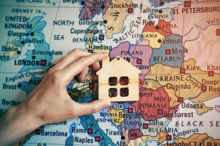 A left hand holding a small wooden house figure with a map in the background
